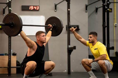 Man lifting weights under coach supervision in a gym setting, focusing on strength training.