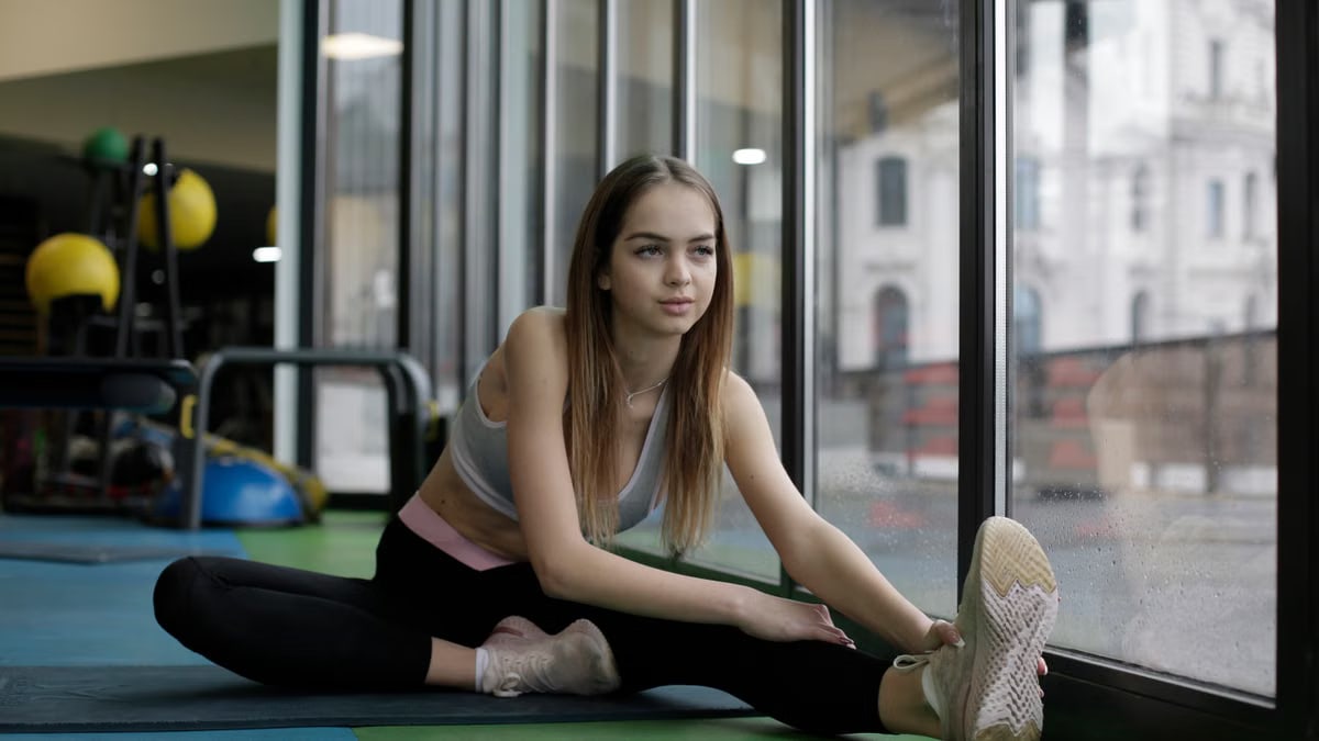 A young woman stretches inside a gym near a large window, emphasizing fitness and wellness.